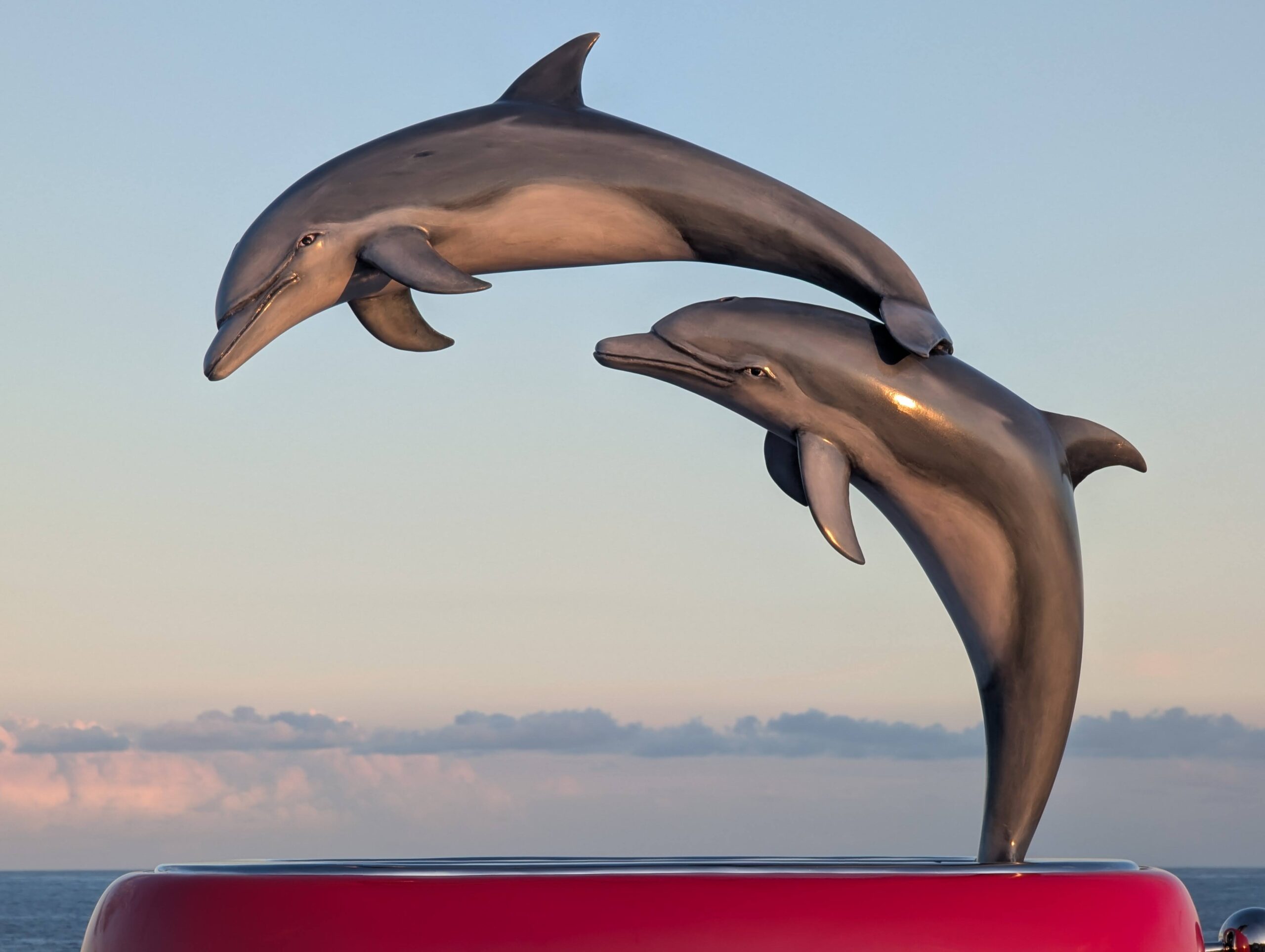 open air artwork of two dolphins jumping in parallel from a red metal box publicly displayed at an exhibition alongside sydney's oceanside suburbs